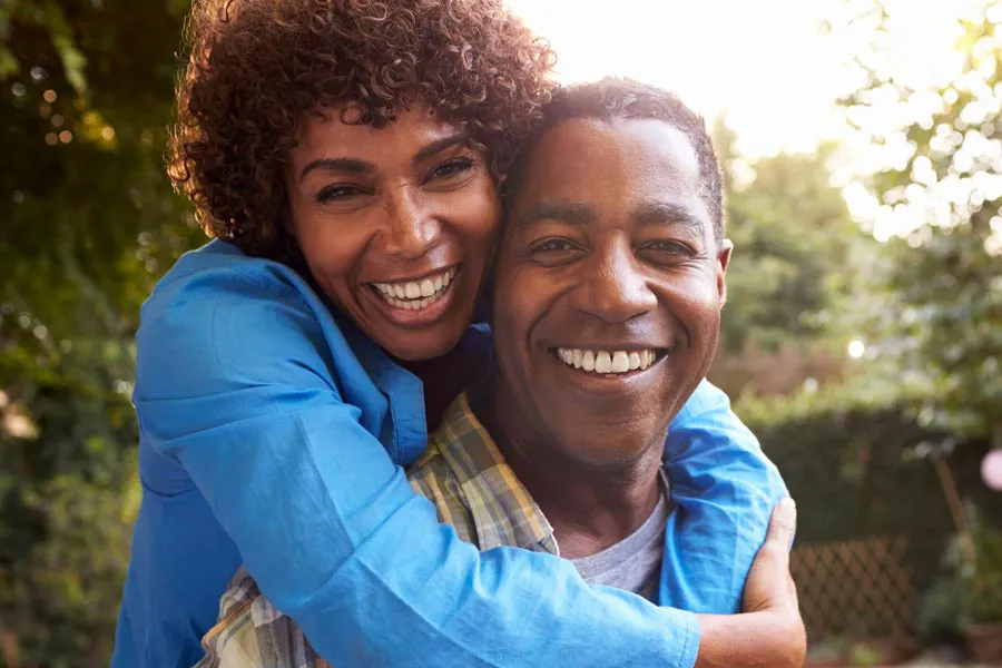 Autoimmune-Doctor A middle-aged African American couple outside, the woman embracing the man from behind pressing her cheek to his, both are smiling due to successful Autoimmune Disorder treatment from Better Life Carolinas of Charlotte in Charlotte Metro.