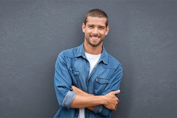 Hormones-for-Men-Doctor A man in a denim shirt stands smiling against a gray-blue wall, pleased with his testosterone hormone therapy from Better Life Carolinas of Charlotte in Charlotte Metro.