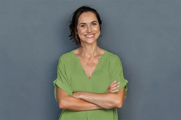 Hormones-for-Women-Doctor A woman in a green shirt stands smiling against a gray-blue wall, pleased with her hormone therapy from Better Life Carolinas of Charlotte in Charlotte Metro.