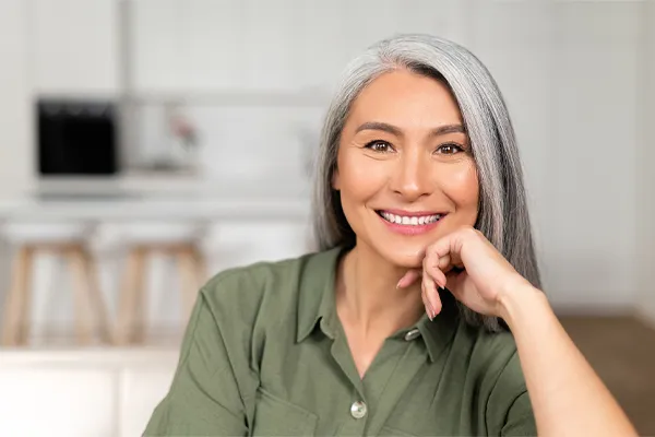 Menopause-Clinic An asian woman with long gray hair and a green shirt sitting and smiling on her couch, happy with her menopause care from [NAME} in Charlotte Metro.