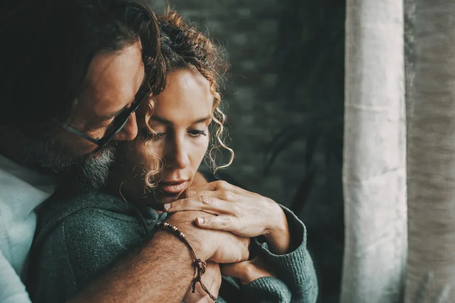 Neurodegenerative-Disorders-Therapy A man embraces a woman from behind in a dimly lit room, their somber expressions showing concerns about a neurodegenerative disease they will get treated by Better Life Carolinas of Charlotte in Charlotte Metro.