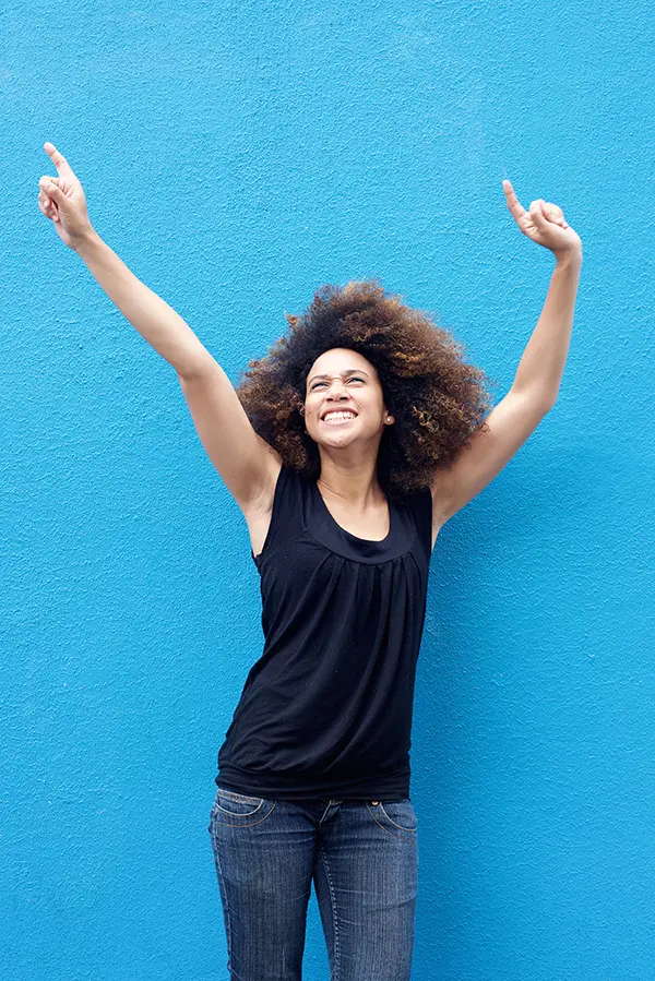 PMS-Treatment A woman in a dark blue tank top standing in front of a bright blue wall, raising her arms in celebration of relief from PMS from Better Life Carolinas of Charlotte in Charlotte Metro.