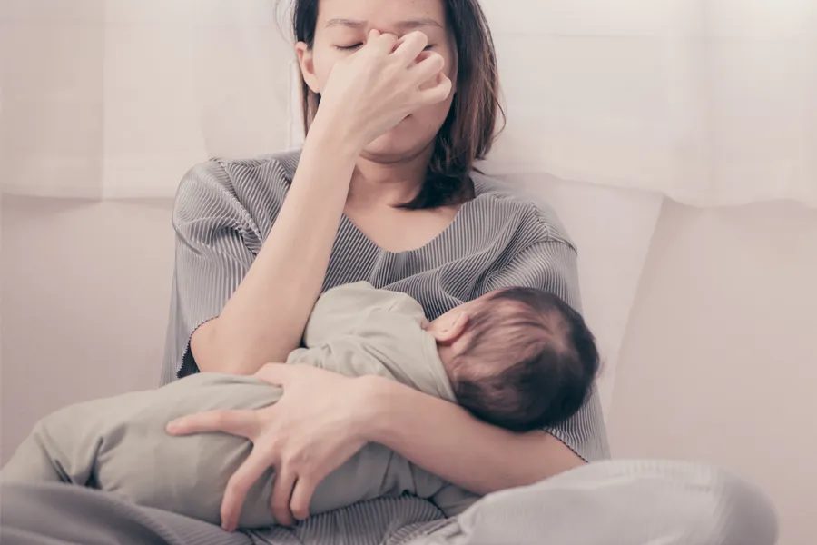 Postpartum-Hormone-Imbalance-Clinic A woman nursing an infant on couch rubs her eyes with exhaustion. Get treatment for postpartum hormone imbalance from Better Life Carolinas of Charlotte in Charlotte Metro.